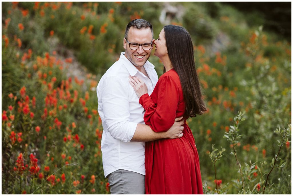 couple kissing in the grassy field with red flowers that match the womans dress