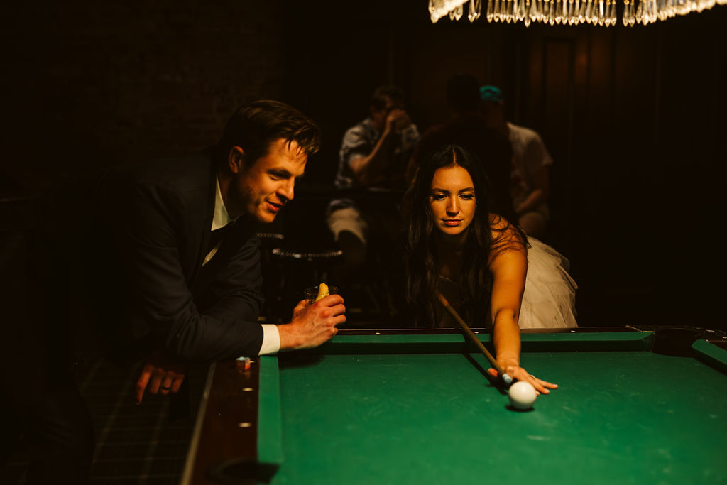 bride and groom play pool in dark bar during elopement