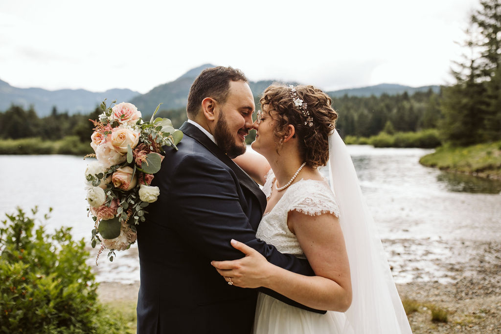 bride and groom smile at each other while standing in front of lake