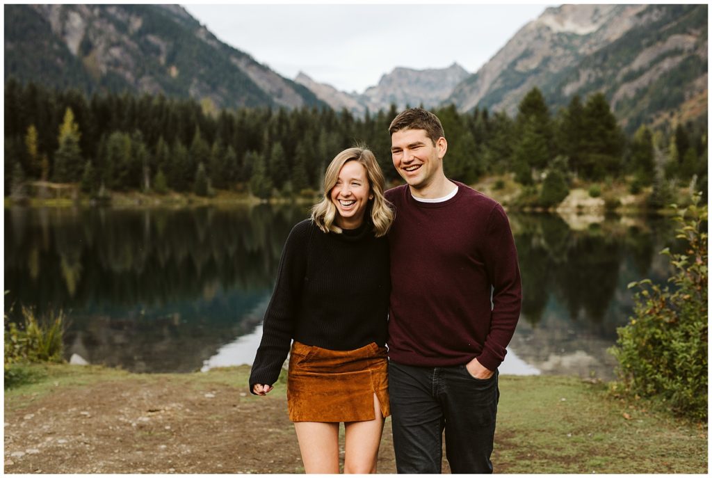 laughing couple fall engagement shoot maroon sweater brown skirt lake reflecting mountains 
