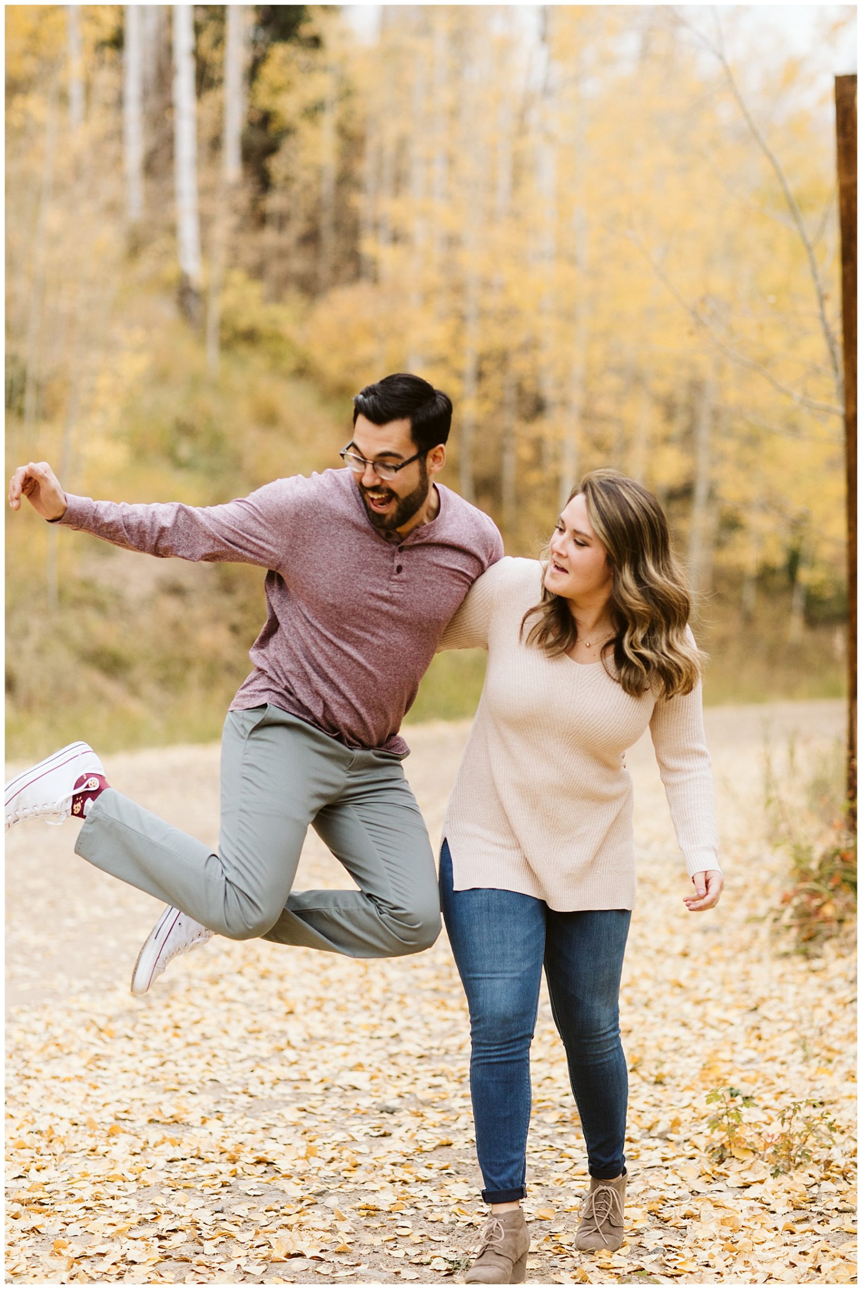 man jumping in air with woman standing next to him laughing at their fall engagement photos.