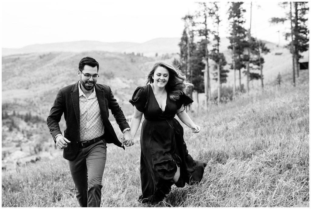 black and white photo of couple walking in grassy field with pine trees in background