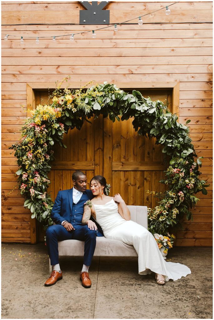 black groom and asian bride sitting underneath a moon shaped floral arch smiling