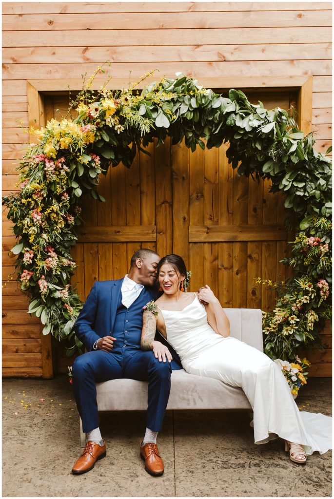 black groom and asian couple sitting underneath a moon shaped floral arch laughing
