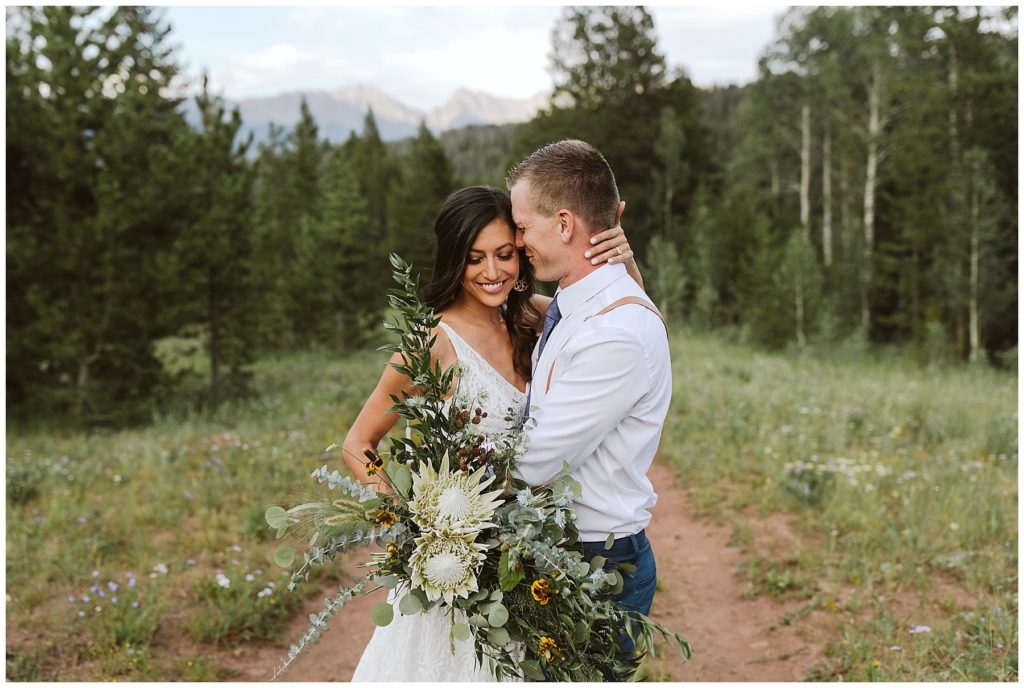brunette woman holding her husbands head to her forehead with a large greenery wedding bouquet