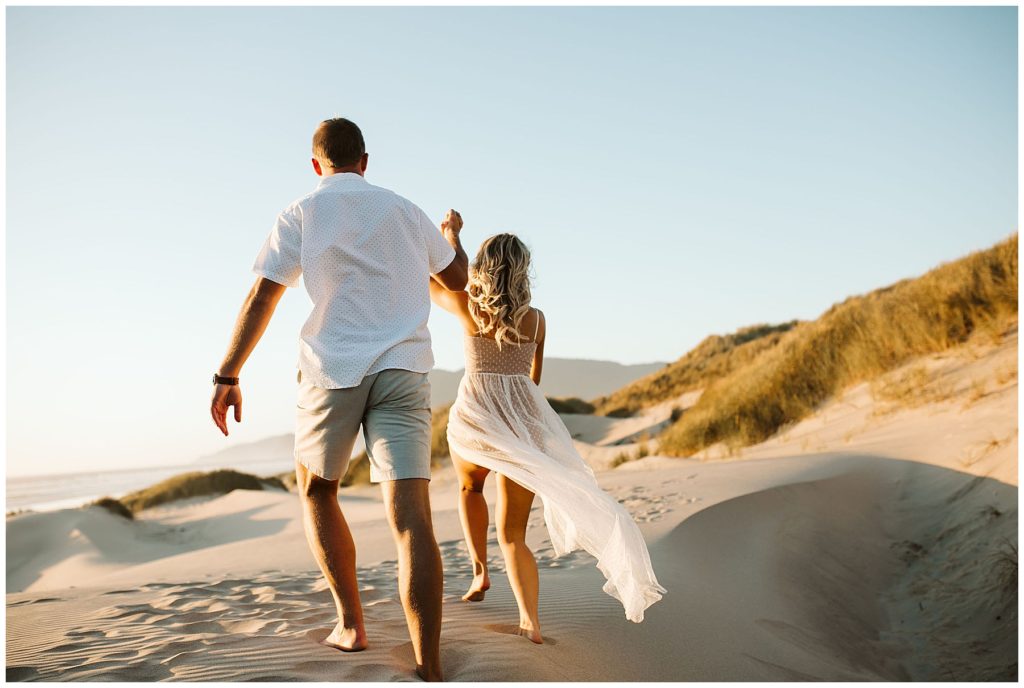 white female and white male holding hands in the air while walking on a beach with their backs facing the camera