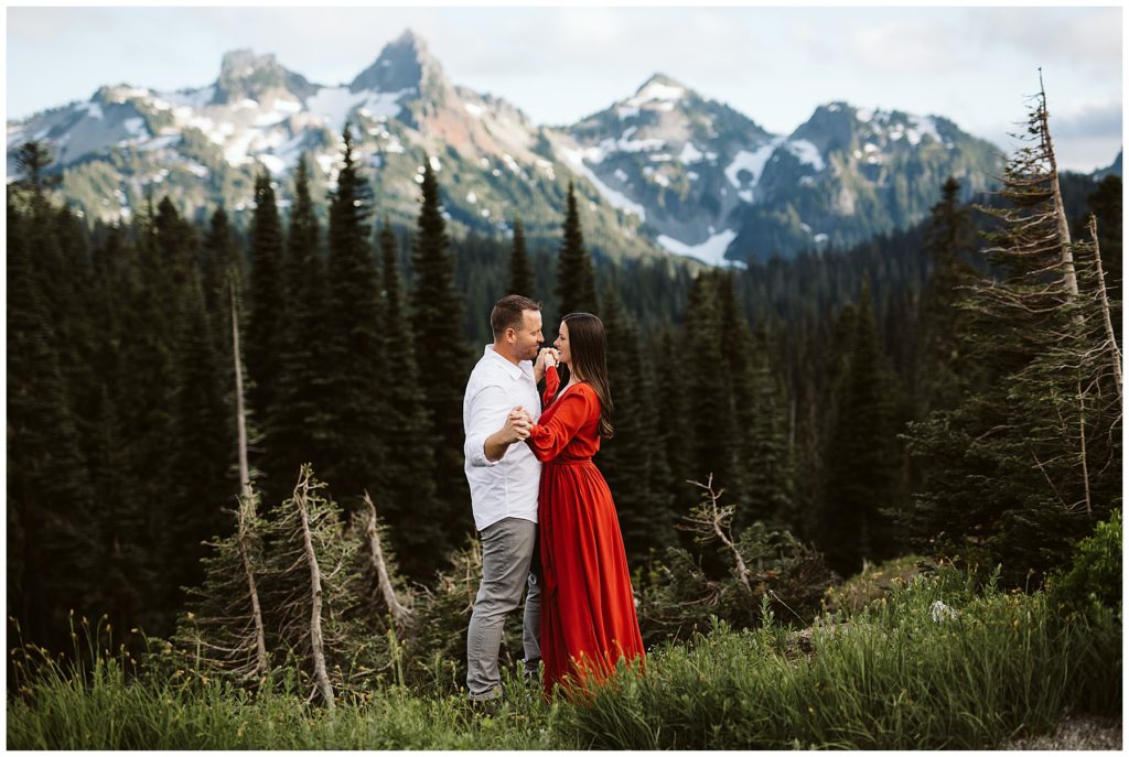 woman standing next to her husband in a striking red dress with the mountains and forest behind them shot by kelly lemon photography 