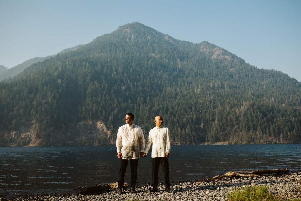 groom and groom stand on the bank of a lake holding hands