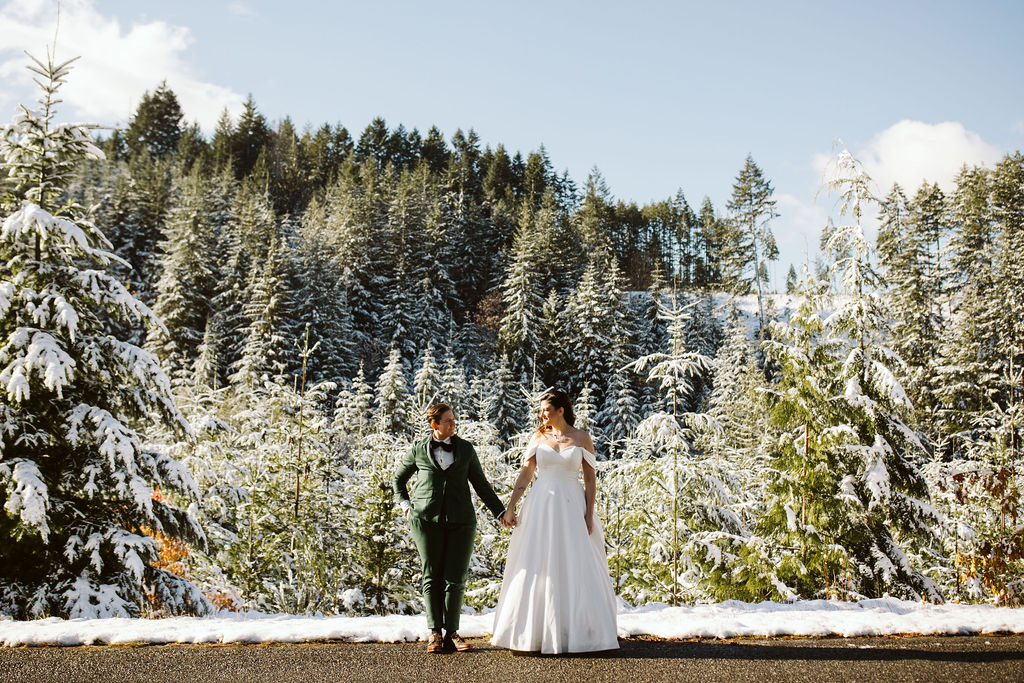 bride in white dress and bride in green suit stand against snowy backdrop