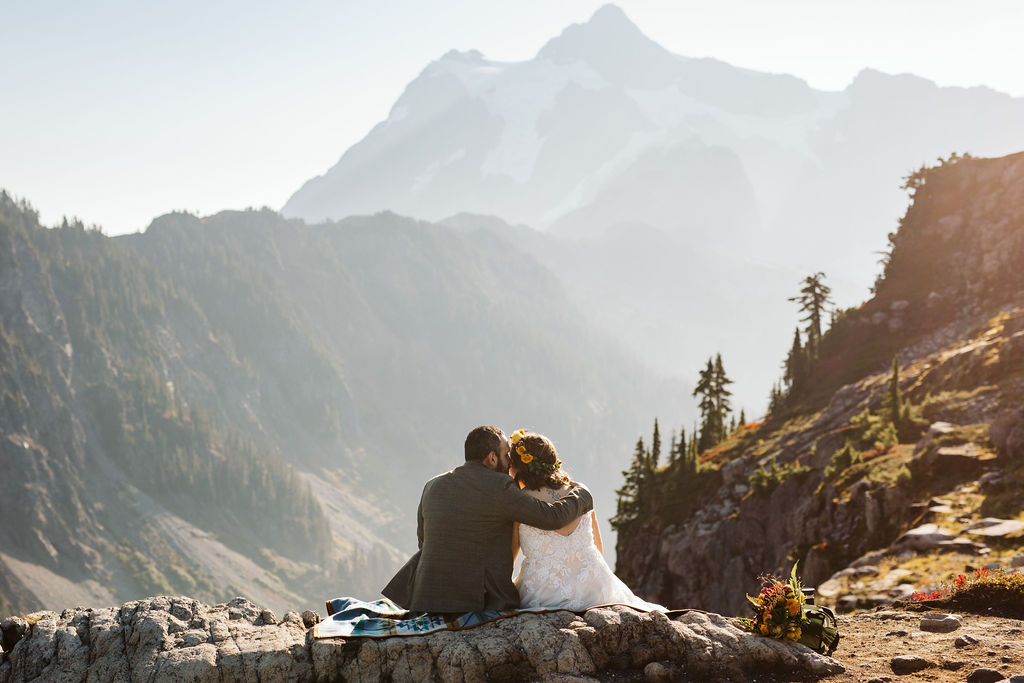 bride and groom sit cuddled on the edge of a cliff during mt baker elopement