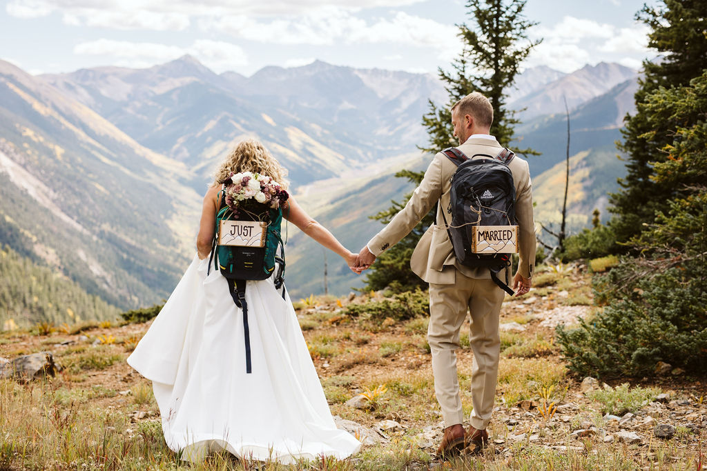 bride and groom wear backpacks and walk towards cliffside