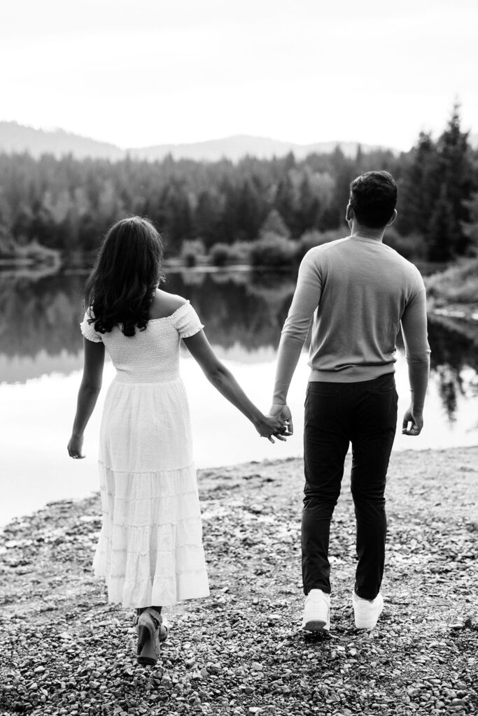 boy and girl hold hands and walk towards lake during engagement session