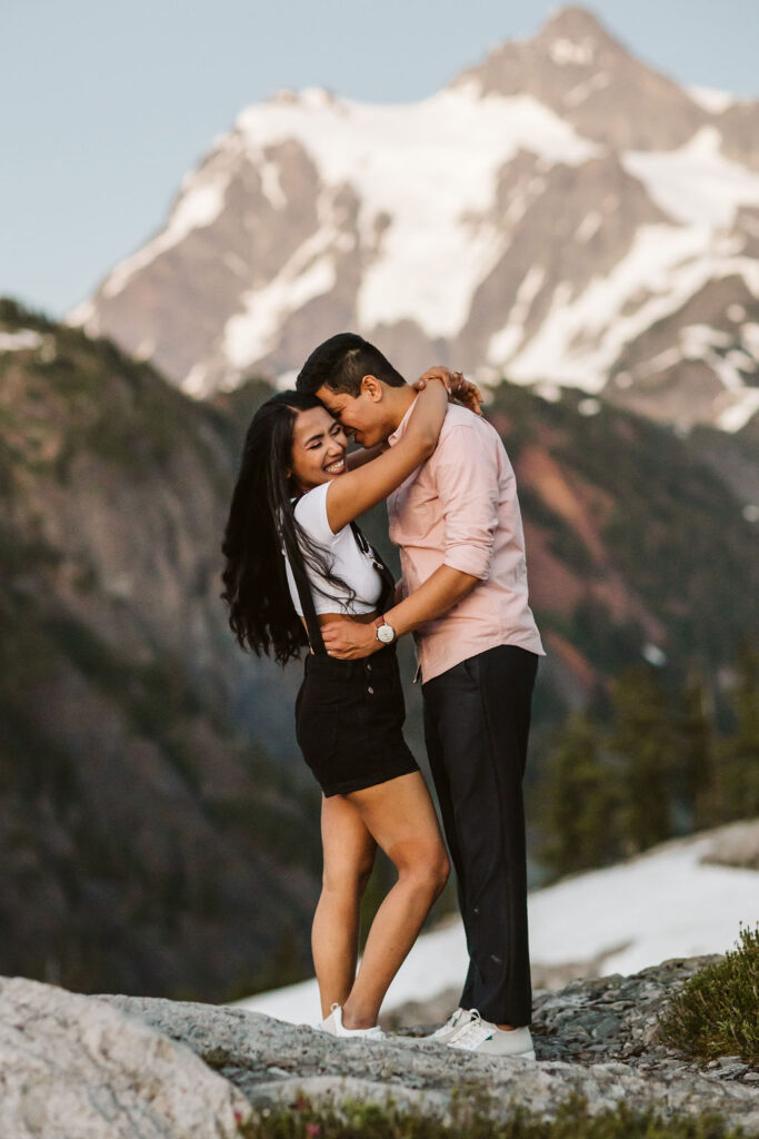 boy and girl nuzzle while snow capped mountains loom behind them during artist point engagement shoot