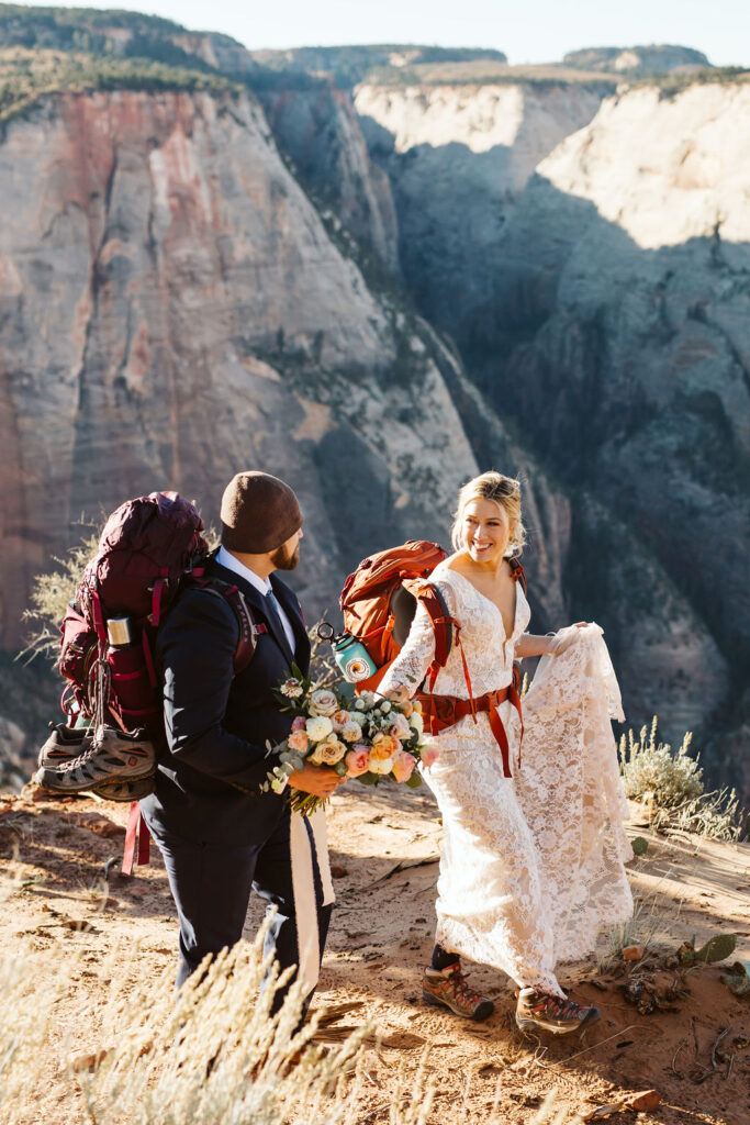 bride and groom wear backpacks and walk along the cliffs during elopement