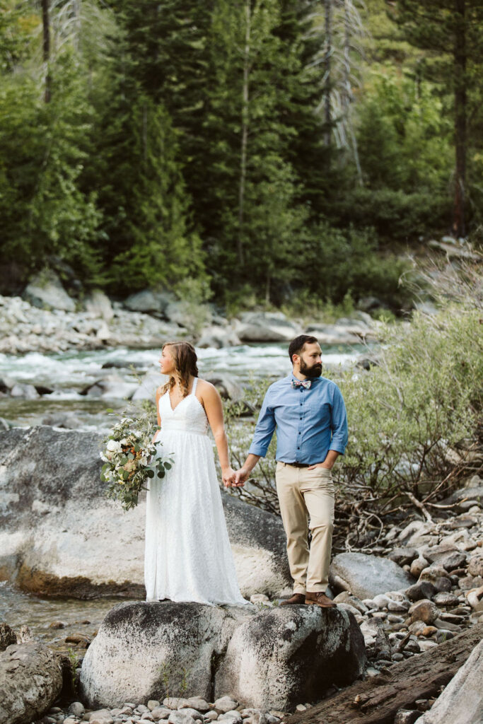 bride and groom hold hands and stand on a rock in a river