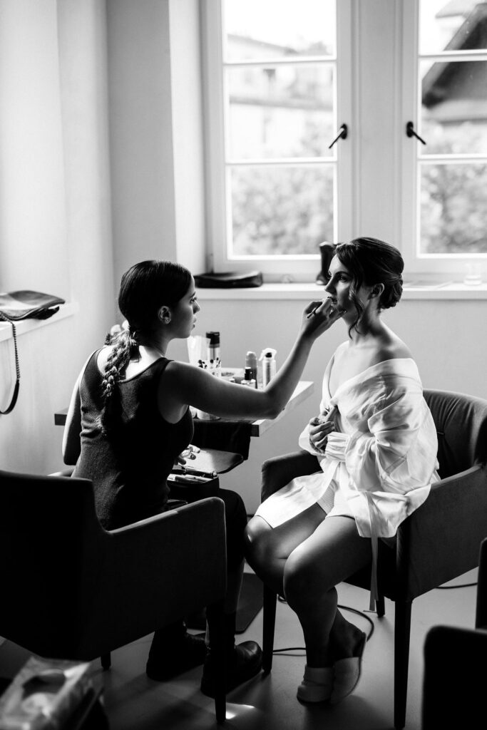 bride sits in window light while someone puts makeup on her
