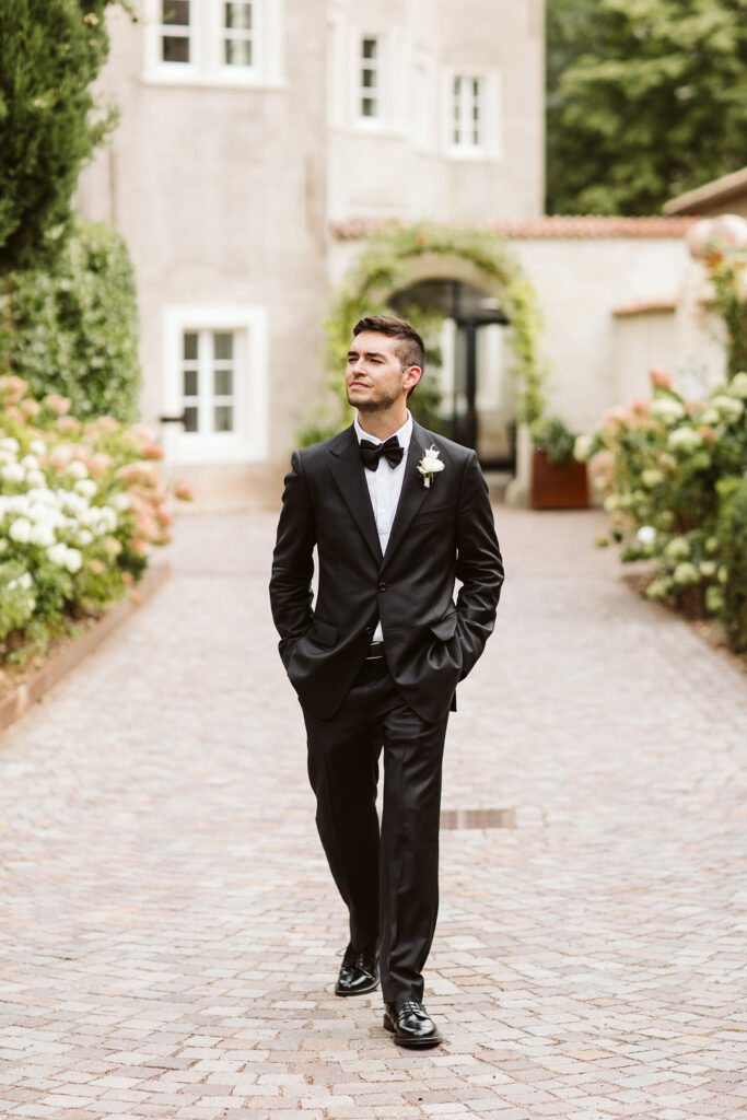 groom walks down cobblestone path, hands in pockets