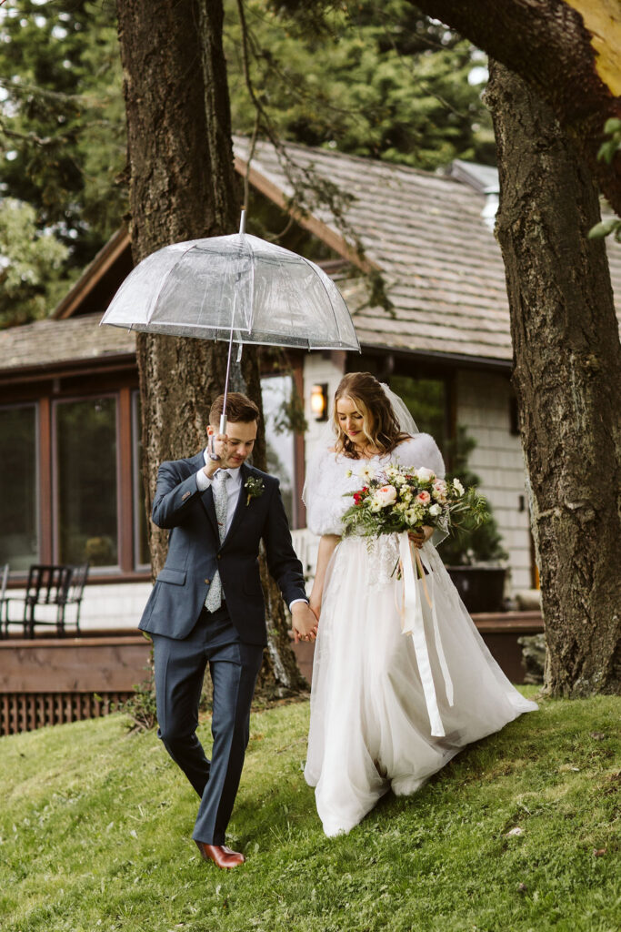 groom helps bride down a grassy hill holding a clear umbrella over them