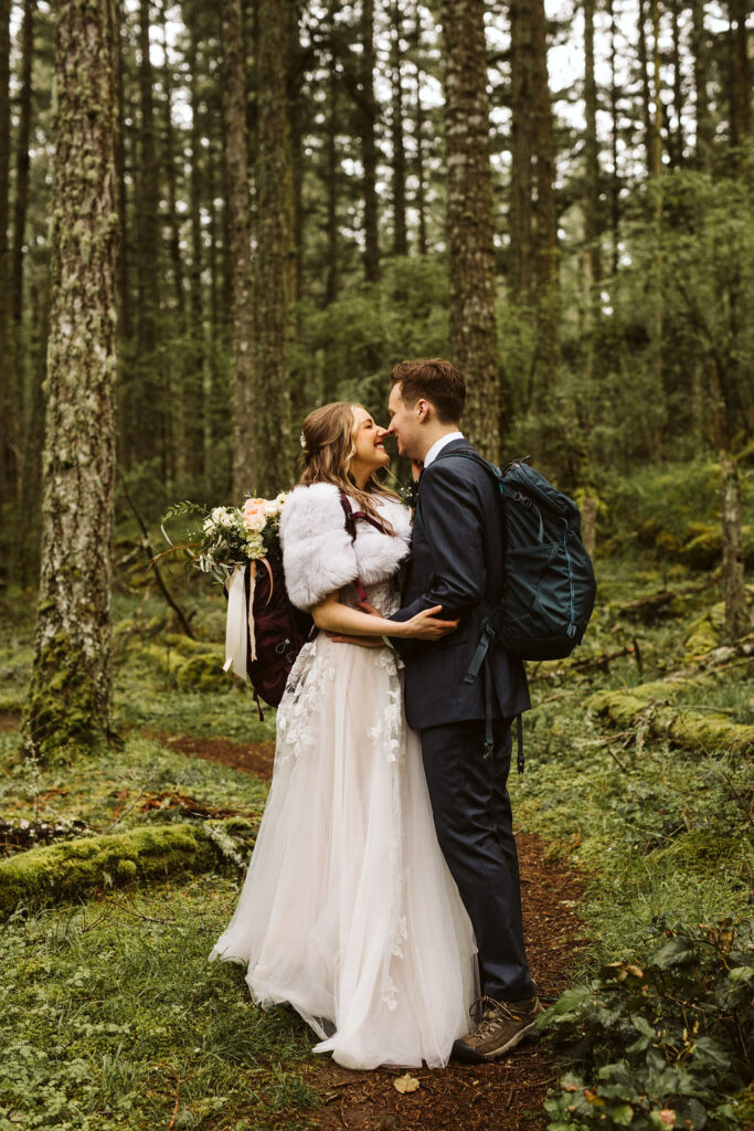 bride and groom face each other in middle of forest while wearing backpacks