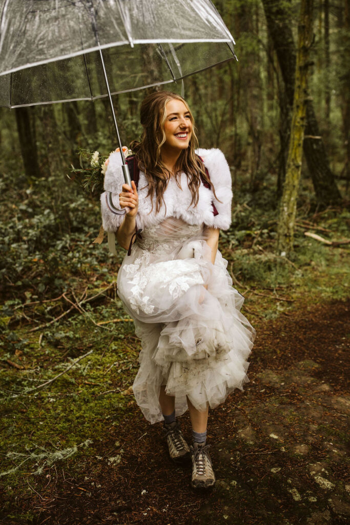 brides smiles and hikes up her dress as she holds clear umbrella in the forest