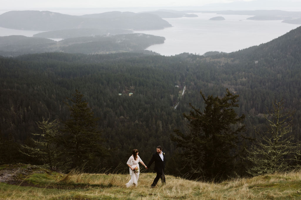 bride and groom walk across a hillside with a view of the lake in the back