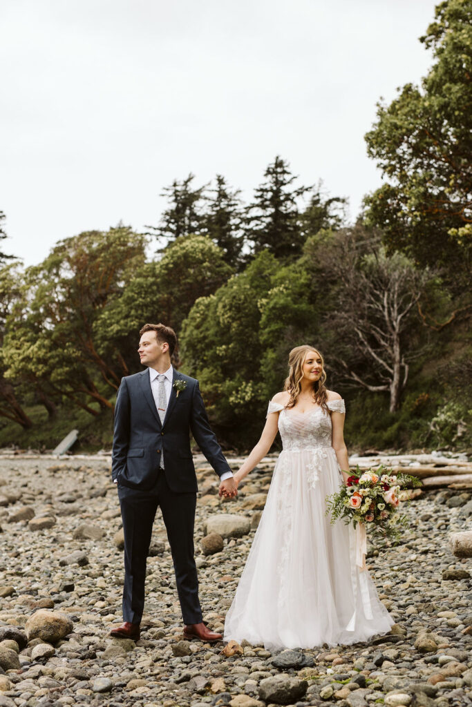 bride and groom stand on rocky beach holding hands and looking in opposite directions