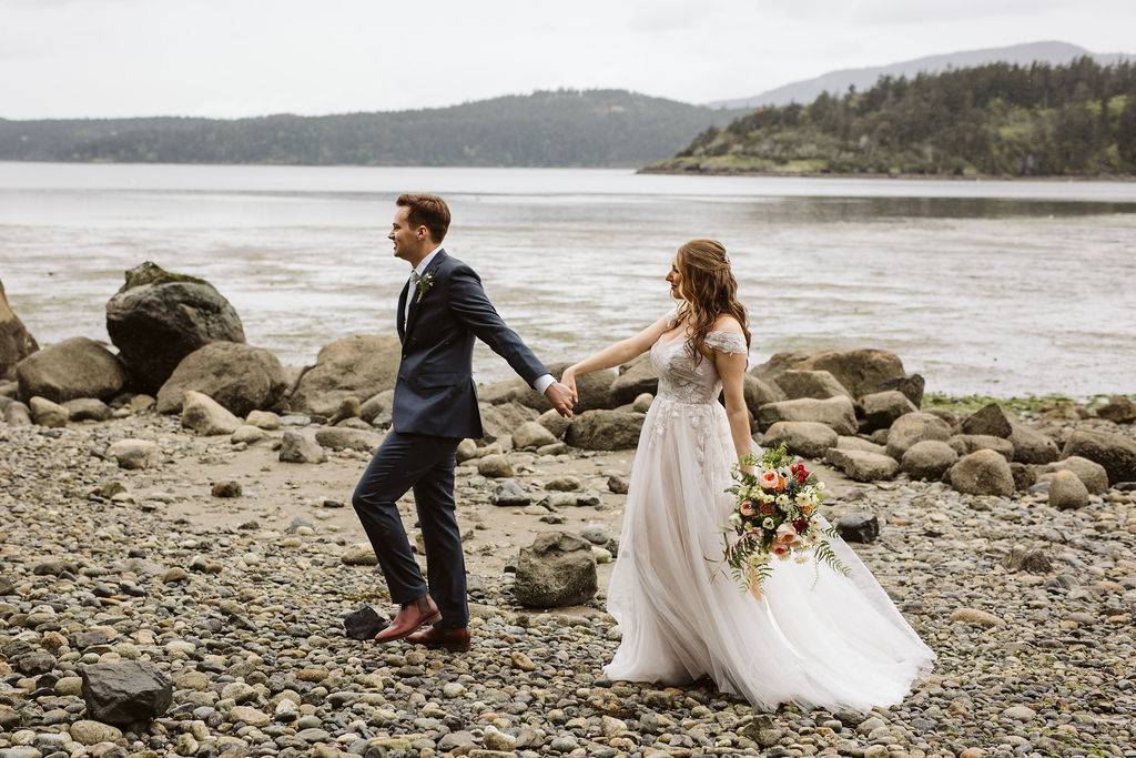 bride and groom walk across rocky beach holding hands