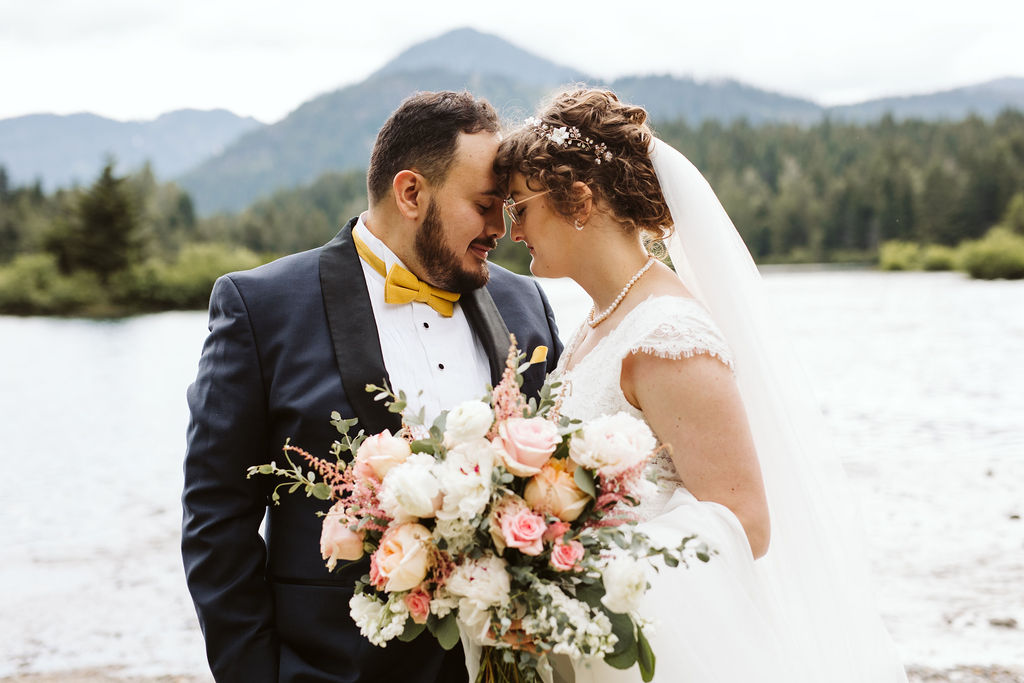 bride and groom stand forehead to forehead in front of lake