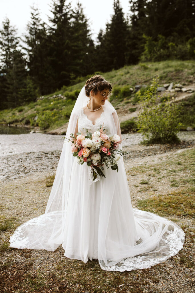 bride stands net to lake looking down at pink and white flowers