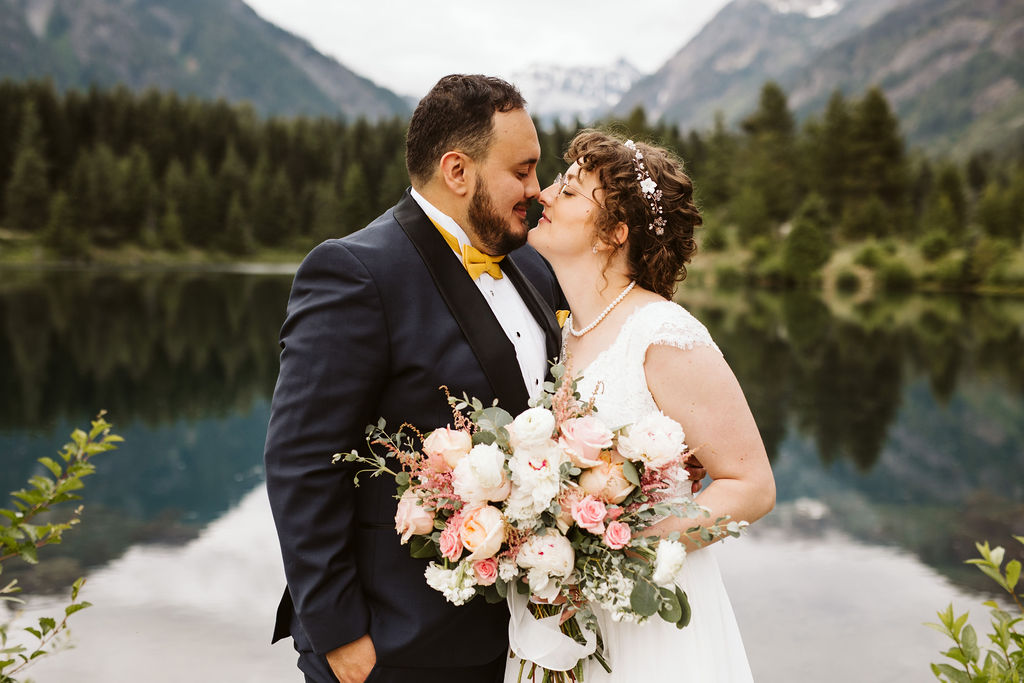 bride and groom stand nose to nose in front of a lake