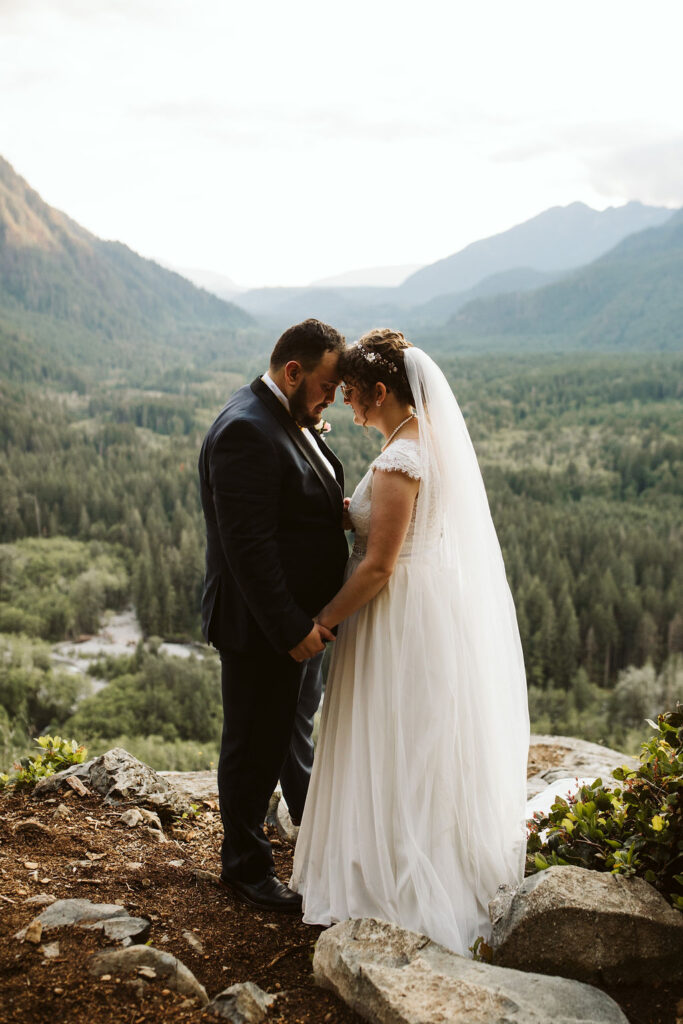 bride and groom stand on the edge of a rock with a view of mountains behind them
