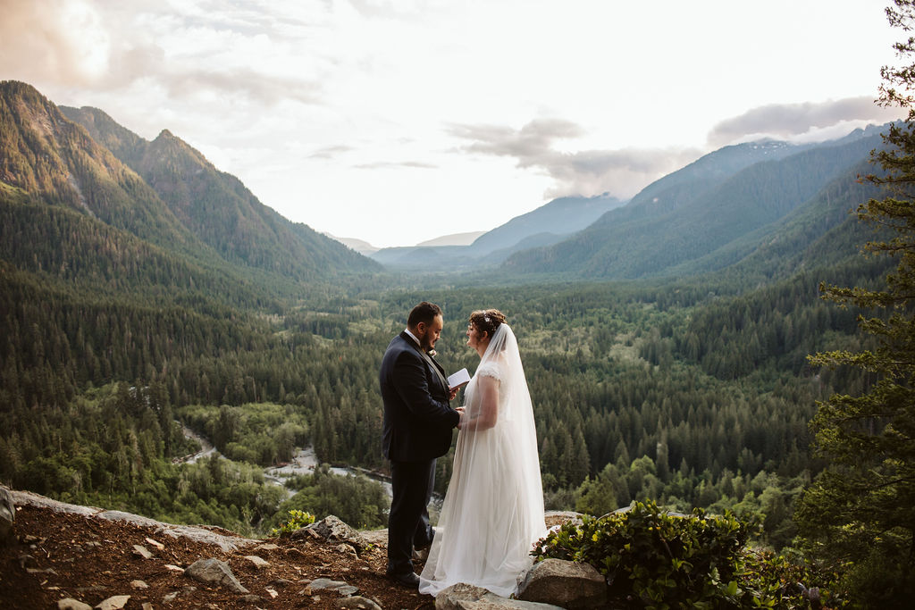 bride and groom stand on the edge of a rock with a view of mountains behind them